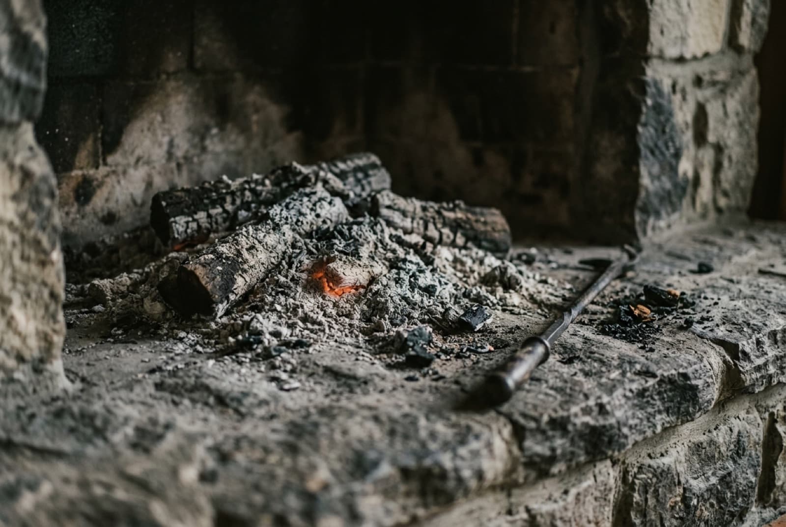Embers nearly smothered under gray ash in a stone fireplace, one patch still glowing
